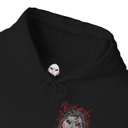 Folded Hooded Sweatshirt of Billy the Puppet staring forward with a sinister grin, his black suit and red bow tie framed by chains, gears, and rusted metal. A dim industrial setting fades behind him as ominous red light highlights his face. The composition emphasizes psychological tension, horror, and the iconic image of the puppet who announces Jigsaw’s deadly games.